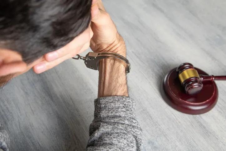 Hombre esposado con la cabeza agachada junto a un mazo de juez sobre una mesa, representando una condena o proceso penal.
