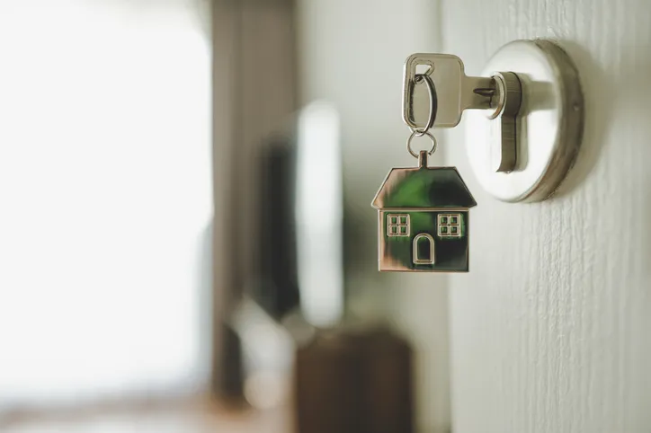 Llave con llavero en forma de casa insertada en la cerradura de una puerta, simbolizando la compra o adquisición de una vivienda.