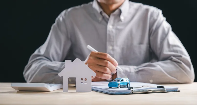 Hombre firmando documentos junto a una calculadora, una figura de casa y un coche de juguete sobre la mesa, representando la gestión de préstamos o hipotecas.