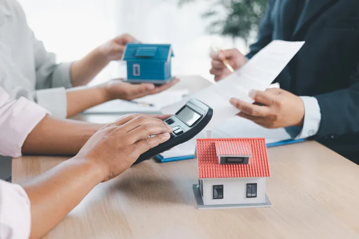 Personas usando una calculadora, revisando documentos y sosteniendo maquetas de casas, representando una gestión hipotecaria o asesoría inmobiliaria.