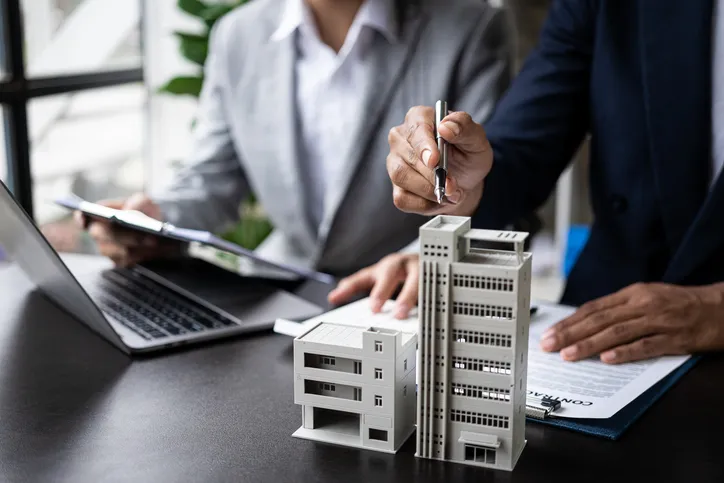 Dos personas firmando un contrato inmobiliario junto a maquetas de edificios, representando una operación de compraventa de propiedad.