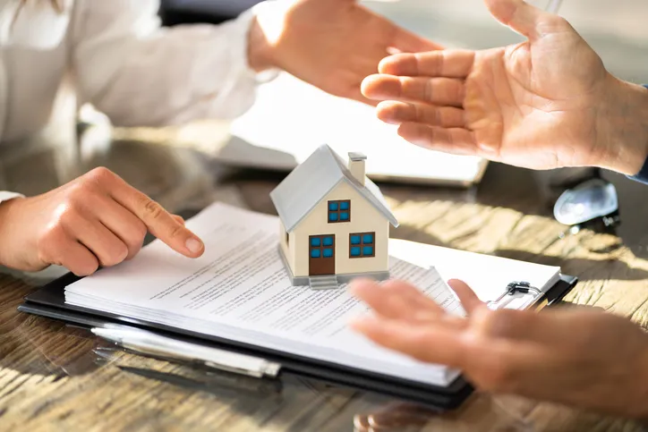 Personas gesticulando y señalando documentos junto a una maqueta de casa, representando una negociación o acuerdo inmobiliario.