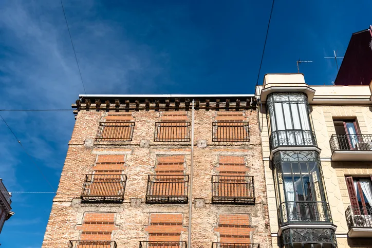 Fachada de un edificio antiguo de ladrillo con balcones enrejados, junto a un edificio moderno con mirador acristalado.