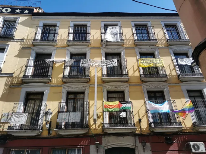 Fachada de un edificio de viviendas con balcones enrejados, del que cuelgan pancartas reivindicativas y banderas de distintos colectivos.