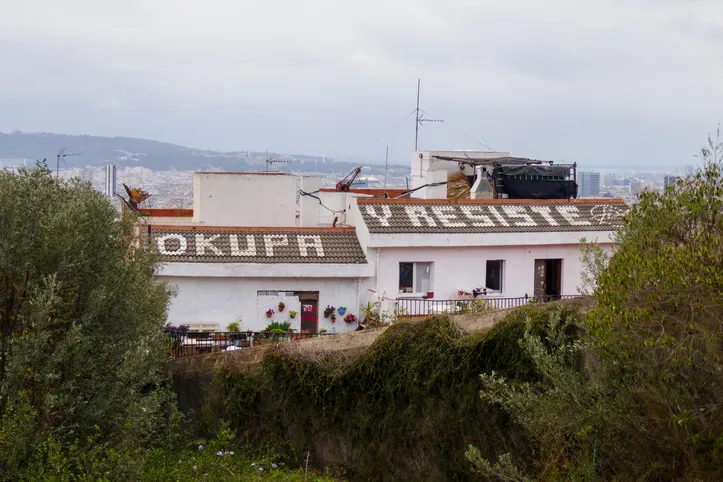 Casas con el texto 'OKUPA Y RESISTE' pintado en el tejado, en una zona con vegetación y vistas a la ciudad.