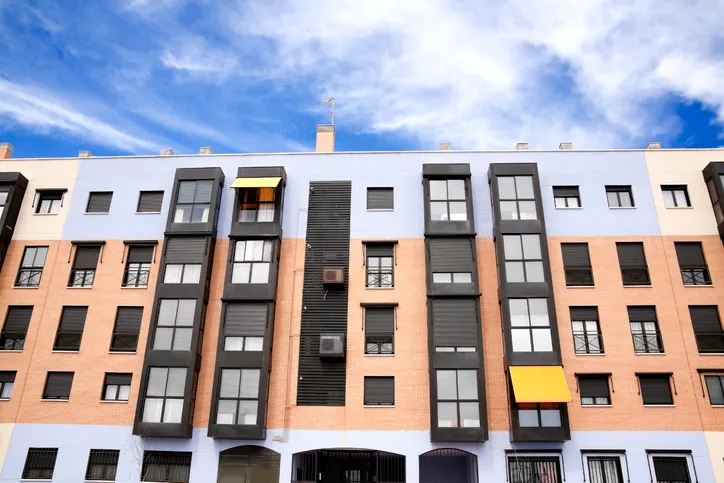 Fachada de un bloque de viviendas en tonos naranja y azul, con balcones y toldos amarillos, bajo un cielo despejado.