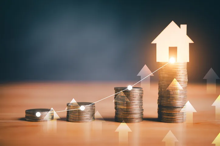 Pilas de monedas en orden ascendente con una silueta de casa iluminada al final, simbolizando el incremento del valor o del coste de la vivienda.