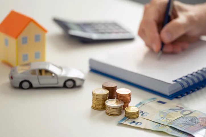 Persona escribiendo en una libreta junto a una calculadora, monedas, billetes de euro y miniaturas de una casa y un coche, representando la planificación de gastos personales.