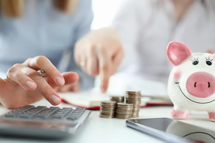 Personas usando una calculadora junto a pilas de monedas y una hucha en forma de cerdito, representando el cálculo de gastos del hogar.