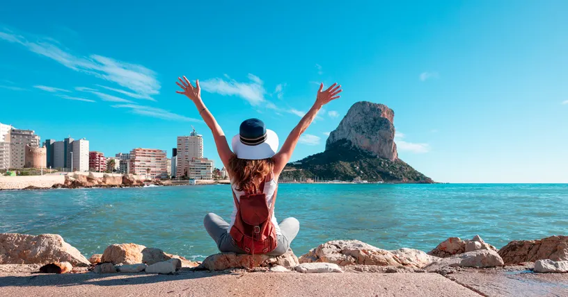 Mujer sentada de espaldas sobre las rocas con los brazos levantados, contemplando el Peñón de Ifach y el skyline de Calpe junto al mar Mediterráneo.