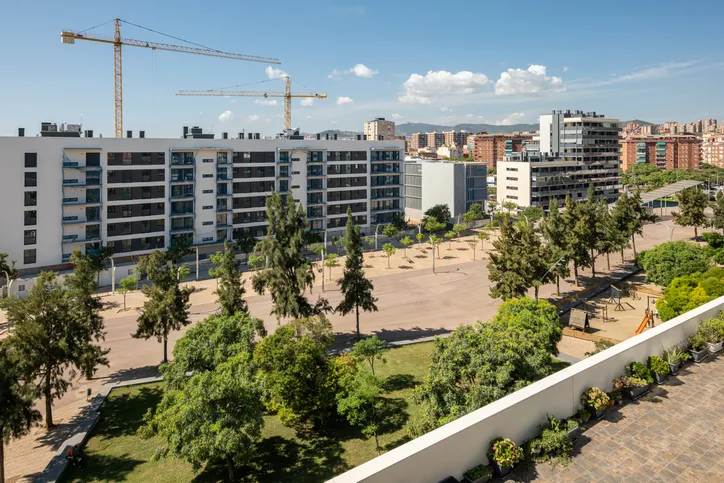 Vista aérea de un nuevo bloque de viviendas en construcción con grúas, rodeado de zonas verdes, parque infantil y edificios residenciales al fondo.