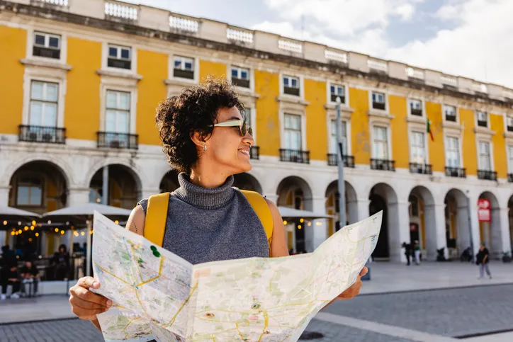 Mujer turista con gafas de sol y mochila consultando un mapa de papel frente a un edificio histórico de fachada amarilla con arcadas.