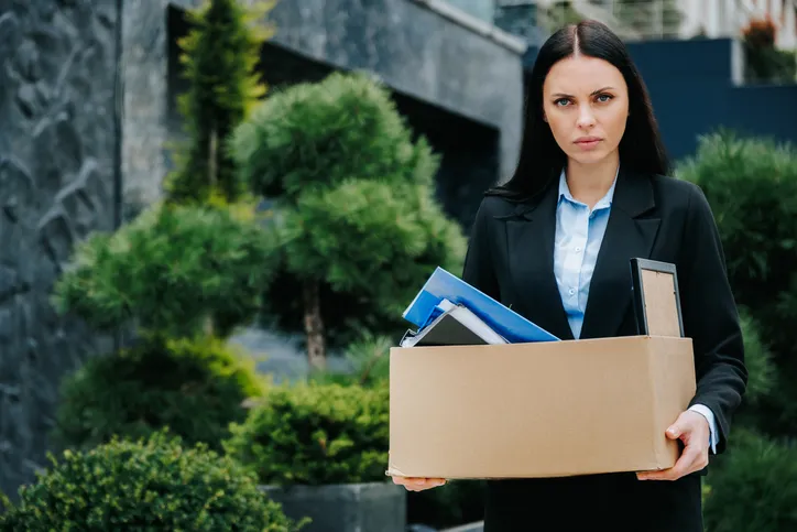 Mujer con traje de oficina y expresión seria sosteniendo una caja de cartón con sus pertenencias tras ser despedida del trabajo.