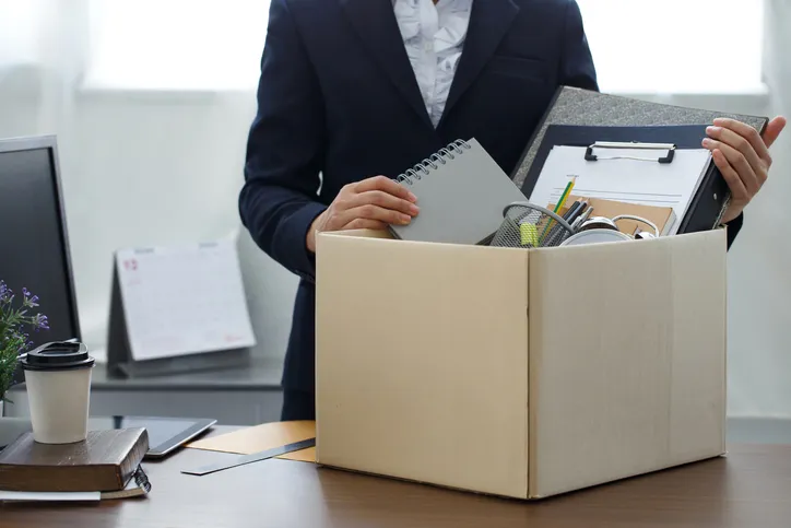 Persona de traje guardando notas, carpeta y objetos personales en una caja de cartón sobre el escritorio de su oficina.