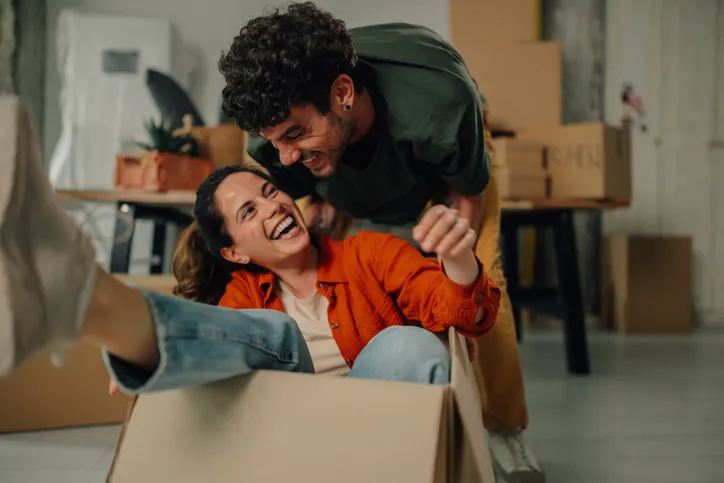 Pareja riendo mientras juega dentro de una caja de cartón en su nuevo hogar, rodeada de cajas de mudanza.