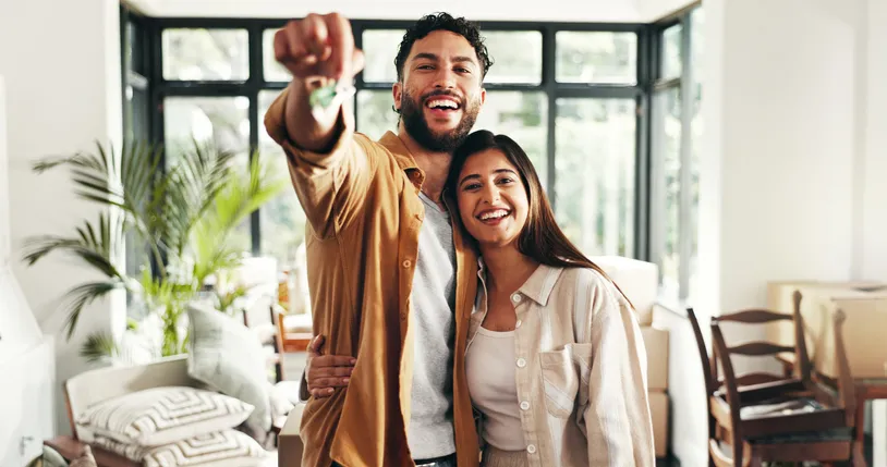 Pareja sonriente mostrando las llaves de su nuevo hogar, abrazados en un salón luminoso rodeado de cajas de mudanza.