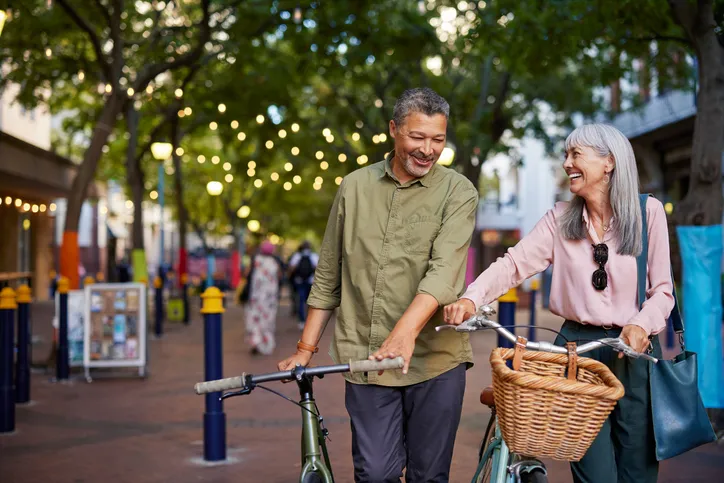 Pareja de personas mayores sonriendo mientras pasean con sus bicicletas por una calle peatonal con luces decorativas.