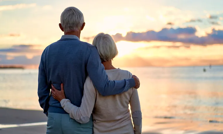Pareja de personas mayores abrazadas de espaldas contemplando el atardecer frente al mar.