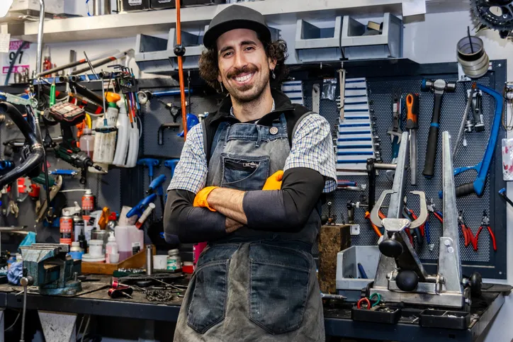 Hombre sonriente con delantal de trabajo, brazos cruzados, en su taller de reparación de bicicletas rodeado de herramientas.
