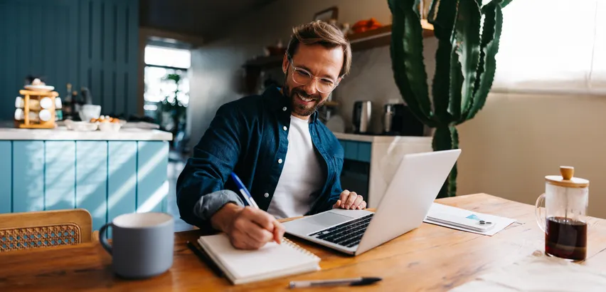 Hombre sonriente tomando notas en un cuaderno mientras trabaja con su portátil en la cocina de casa.