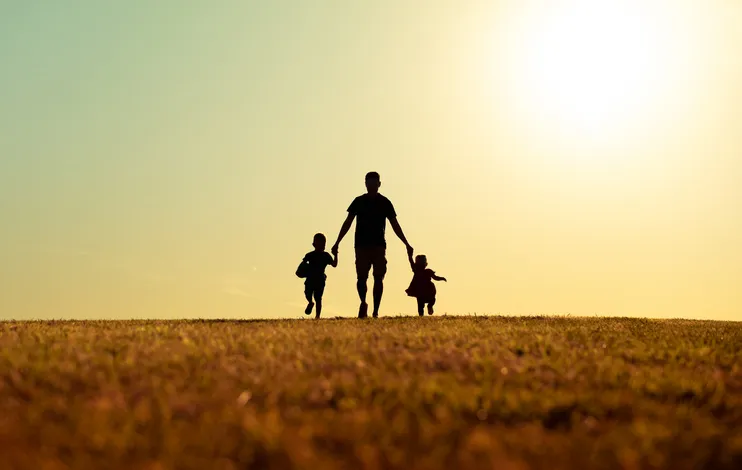 Silueta de un padre caminando de la mano de sus dos hijos por un campo al atardecer.