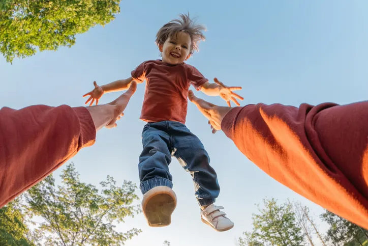 Niño sonriente siendo lanzado al aire por un adulto, visto desde abajo con árboles y cielo de fondo.